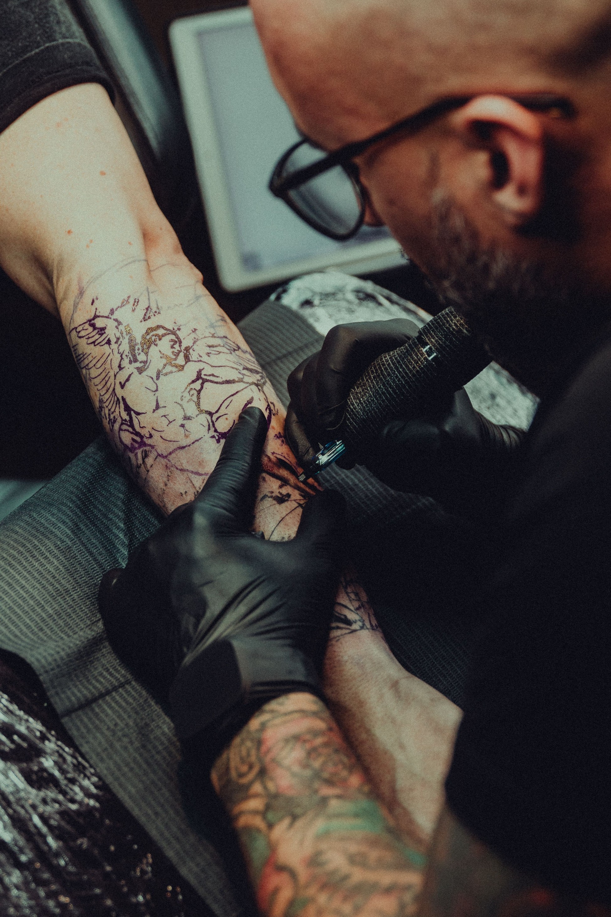 Tattoo artist in a felt hat and dress shirt holding a tattoo machine beside the studio chair, sleeve tattoos on show.