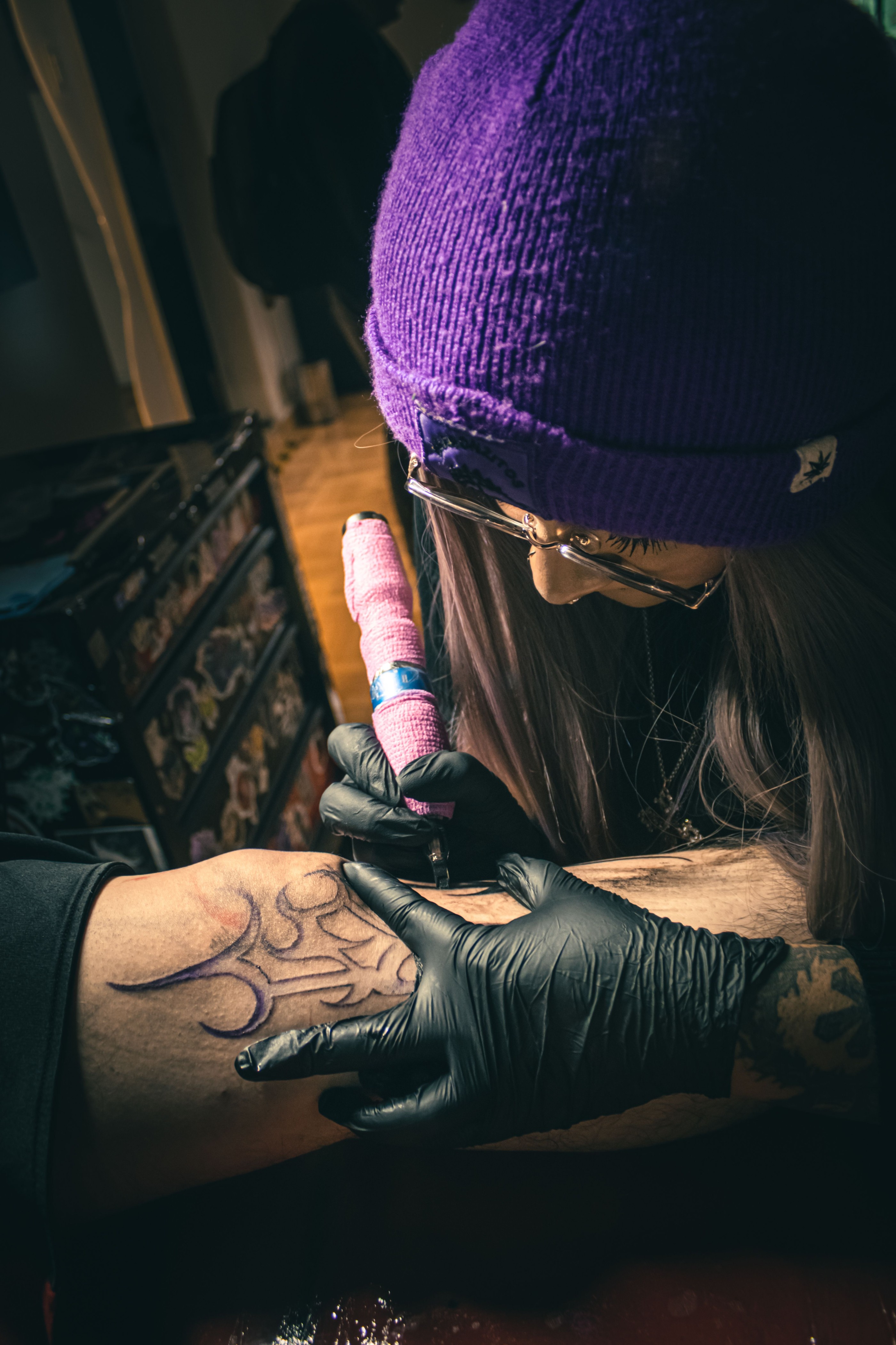 Close-up of a gloved tattoo artist in a purple beanie outlining a design on a client's forearm.